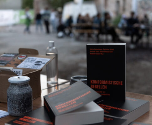 Ein Tisch mit einigen Ausgaben von Konformistische Rebellen, eines der Bücher steht hochkant. Im Hintergrund ein Publikum.