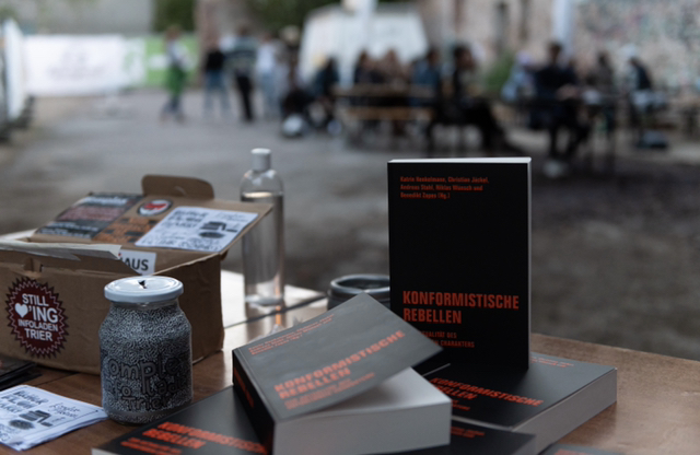 Ein Tisch mit einigen Ausgaben von Konformistische Rebellen, eines der Bücher steht hochkant. Im Hintergrund ein Publikum.